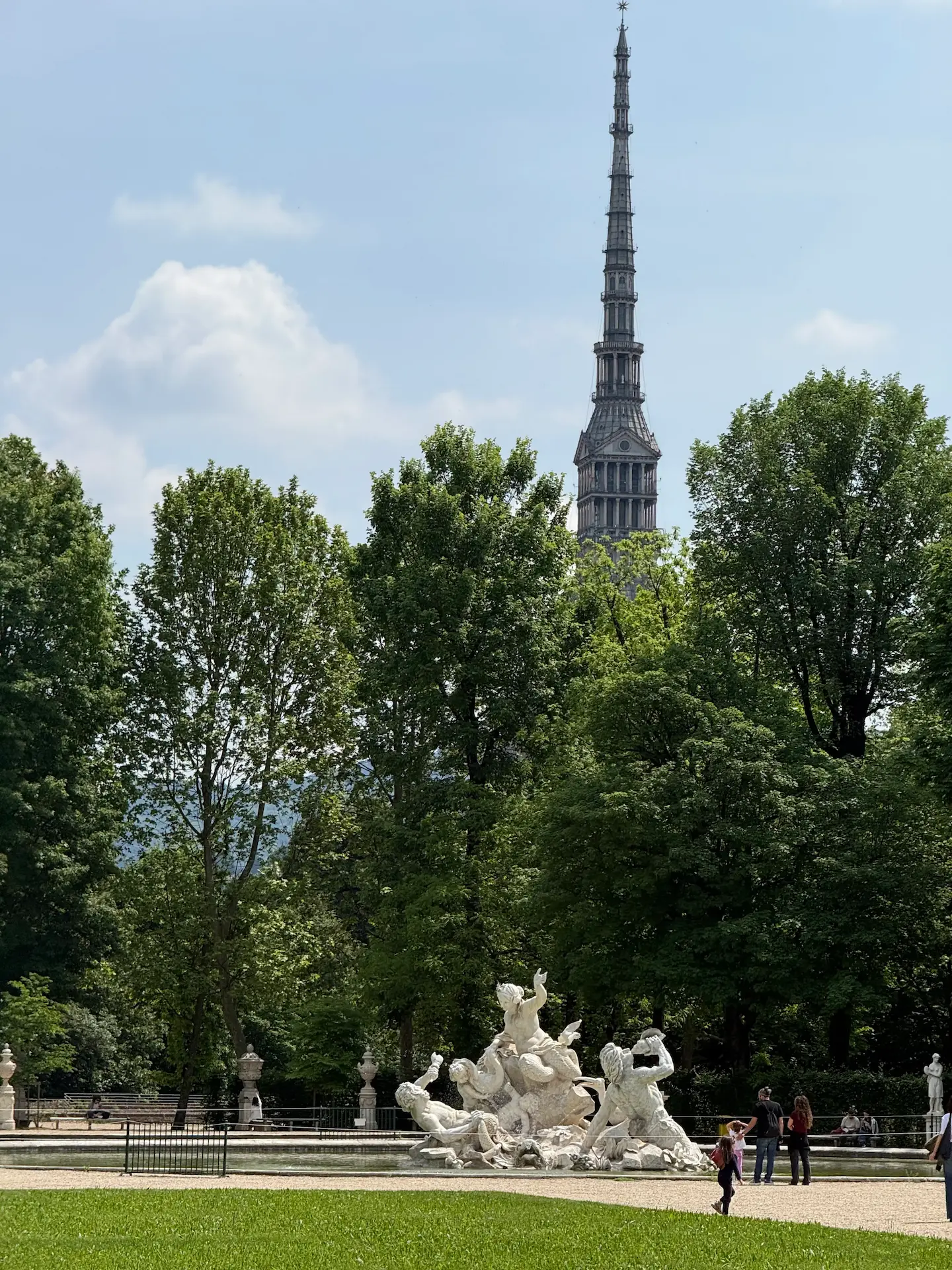 La guglia della Mole Antonelliana di Torino che svetta oltre gli alberi dei Giardini Reali, con la Fontana dei Tritoni in primo piano e alcune persone che passeggiano in una giornata di sole.