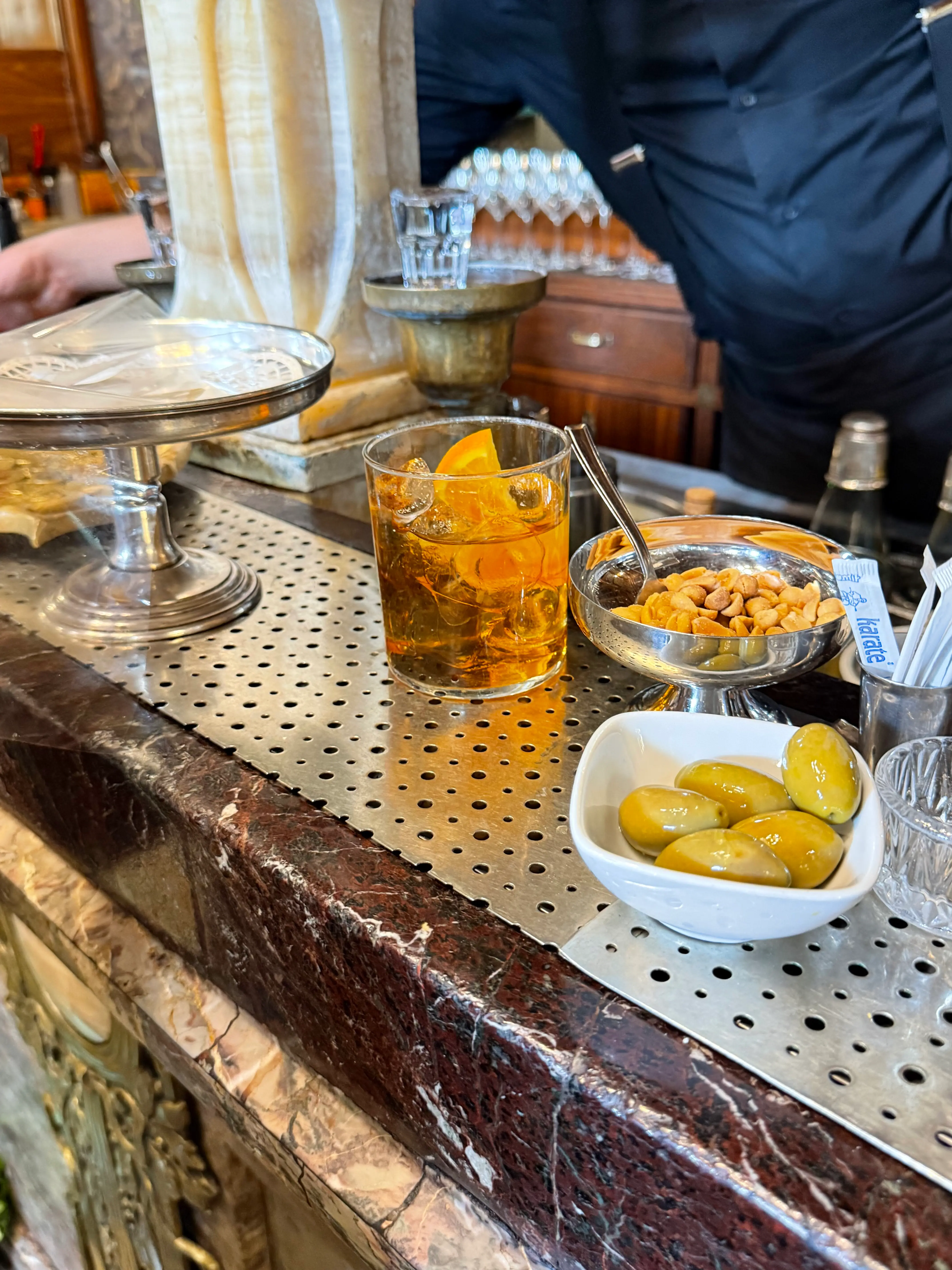 Un bicchiere di Vermut bianco con ghiaccio e una fetta d'arancia servito sul bancone storico del Caffè Mulassano di Torino, accompagnato da piattini con olive verdi e arachidi.