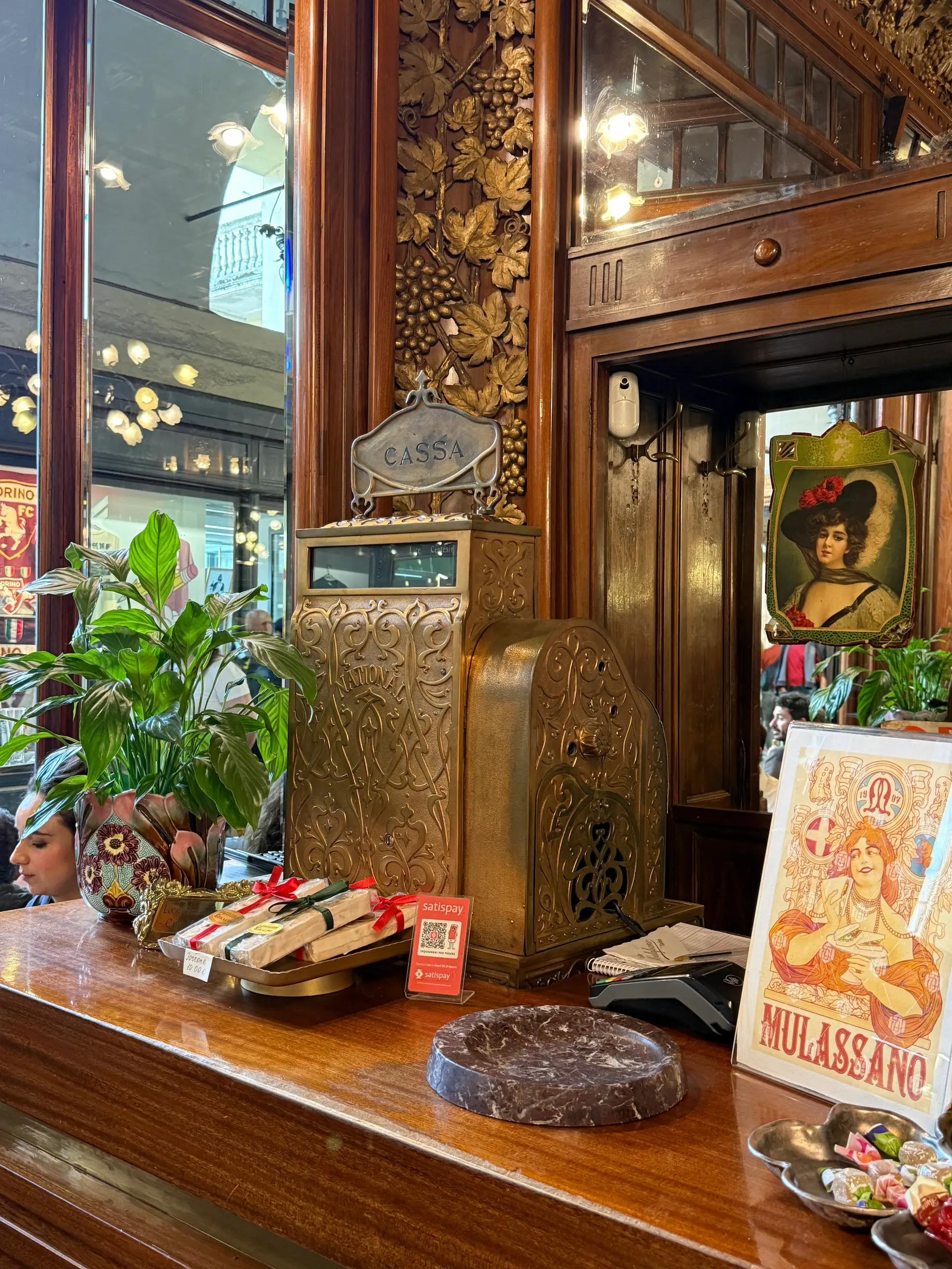 Un registratore di cassa d'epoca in ottone lavorato sul bancone in legno del Caffè Mulassano di Torino, circondato da specchi, boiserie decorate con motivi di foglie e locandine storiche del locale.