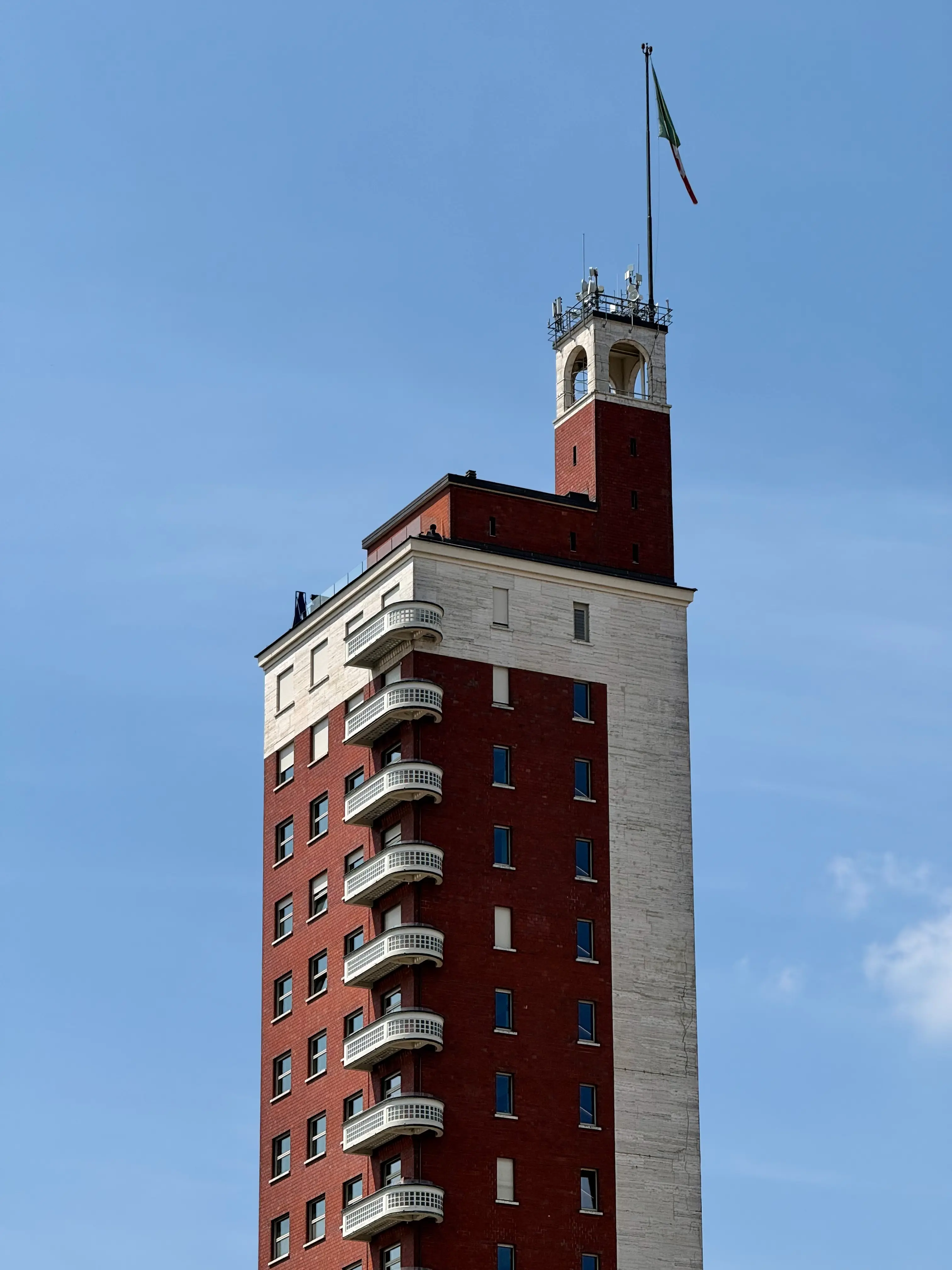 Vista ravvicinata della Torre Littoria, l'edificio razionalista nel centro di Torino, con la sua caratteristica facciata in mattoni rossi e pietra bianca e la colonna di balconi sovrapposti, sullo sfondo di un cielo azzurro limpido e una bandiera italiana in cima.