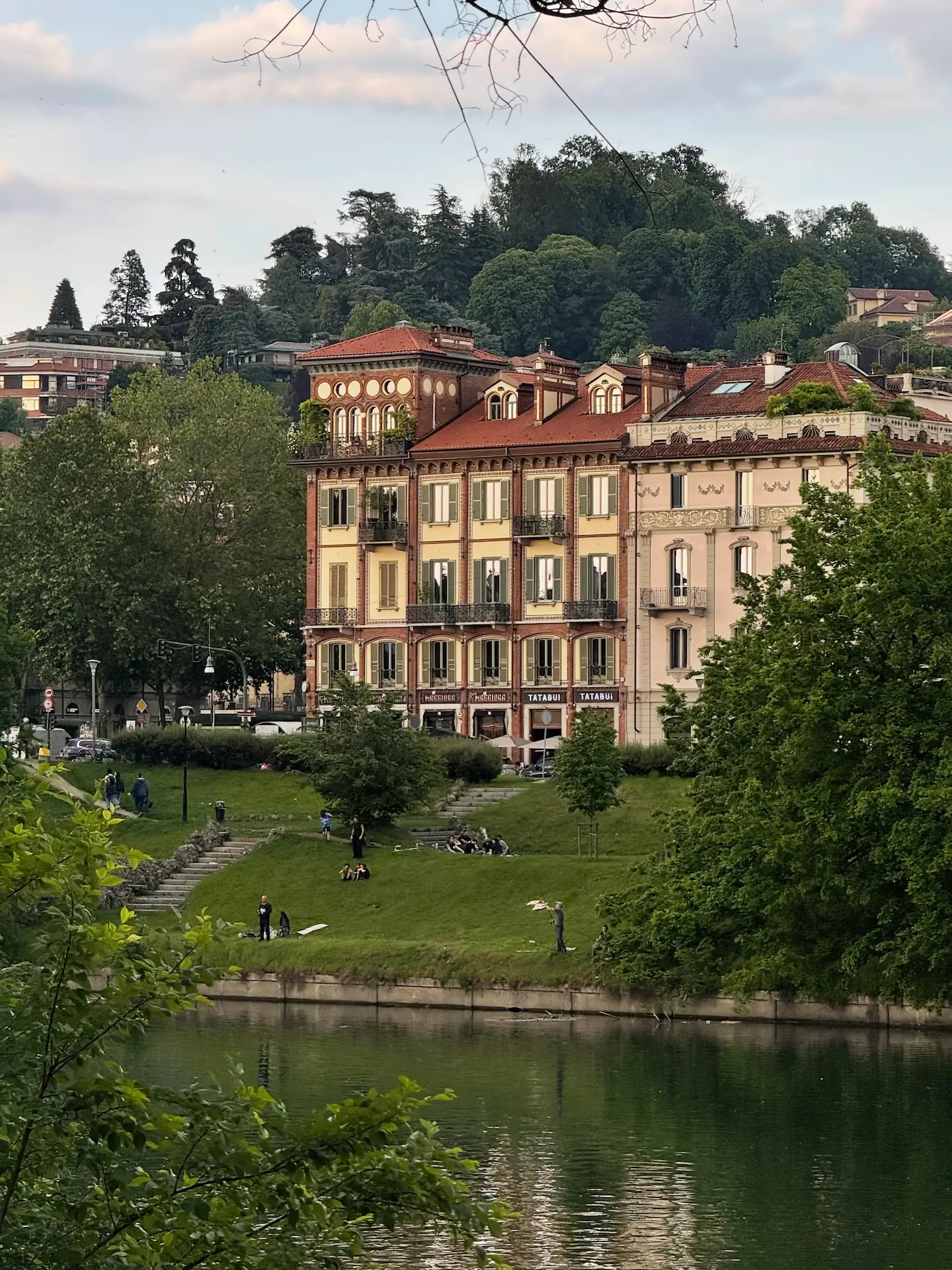 Scorcio del fiume Po a Torino con i Murazzi e le sponde verdi della collina torinese, dove alcune persone si rilassano sul prato davanti a palazzi storici in stile Liberty e alberi rigogliosi.