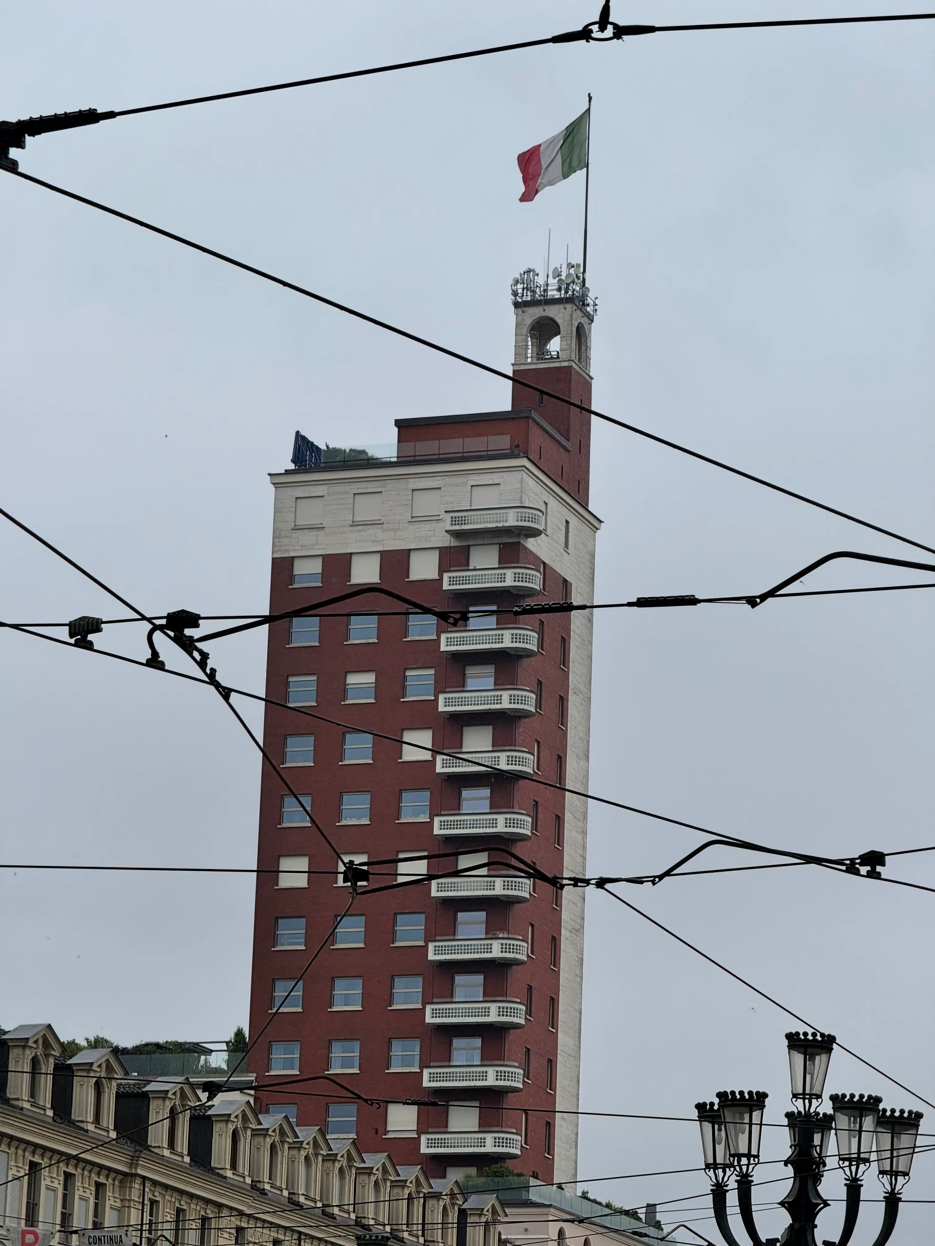 Inquadratura dal basso della Torre Littoria a Torino, incorniciata e parzialmente intersecata dai cavi aerei del tram in primo piano, con la bandiera italiana che sventola sulla sommità dell'edificio in una giornata nuvolosa.