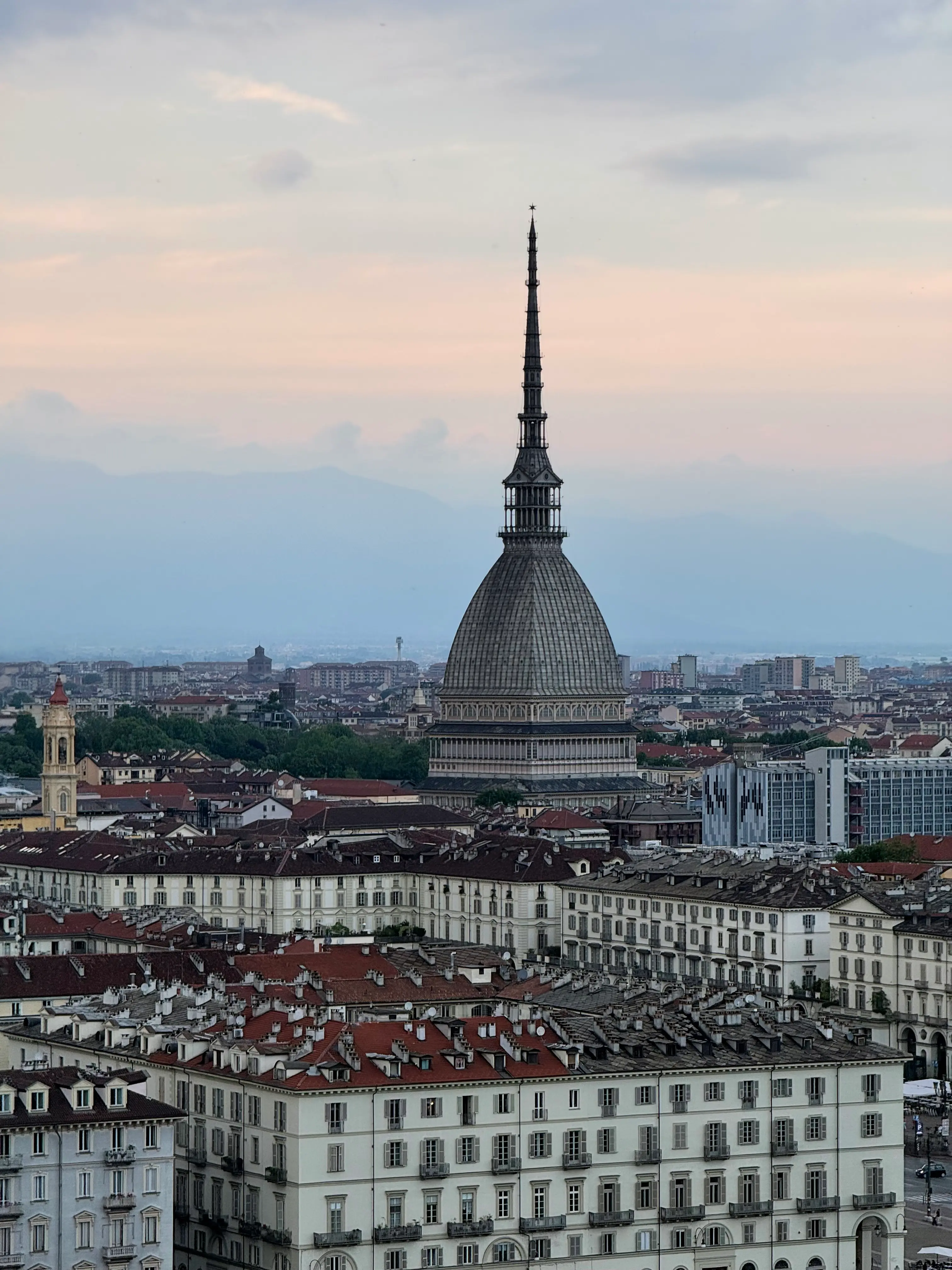 Vista panoramica dall'alto della città di Torino, con la Mole Antonelliana che domina il paesaggio urbano di tetti rossi e facciate chiare, sullo sfondo delle Alpi e di un cielo rosato al tramonto.