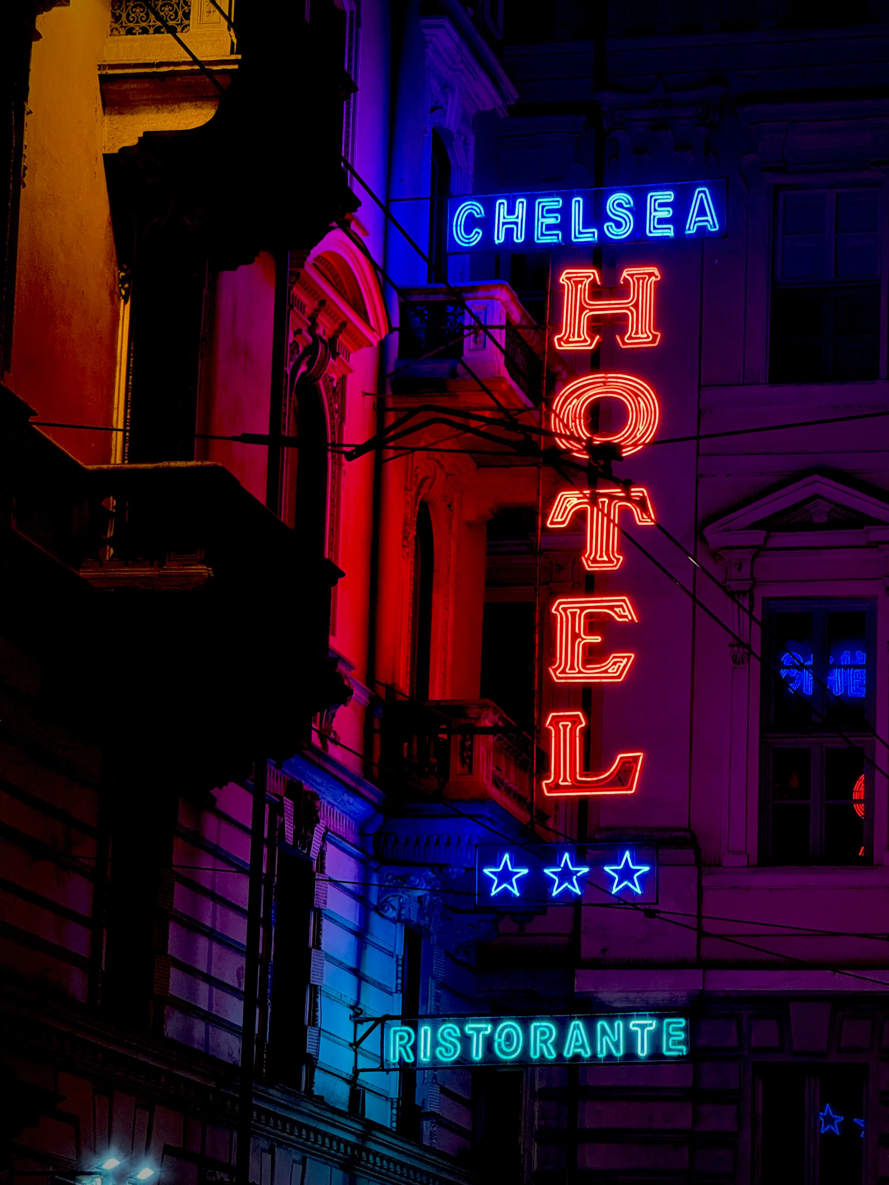 Insegne al neon colorate di un hotel a Torino illuminate di notte; la scritta 'CHELSEA' e tre stelle brillano di blu, la parola 'HOTEL' disposta in verticale è di colore rosso e l'insegna 'RISTORANTE' in basso è di colore azzurro, proiettando riflessi violacei sulla facciata del palazzo storico.