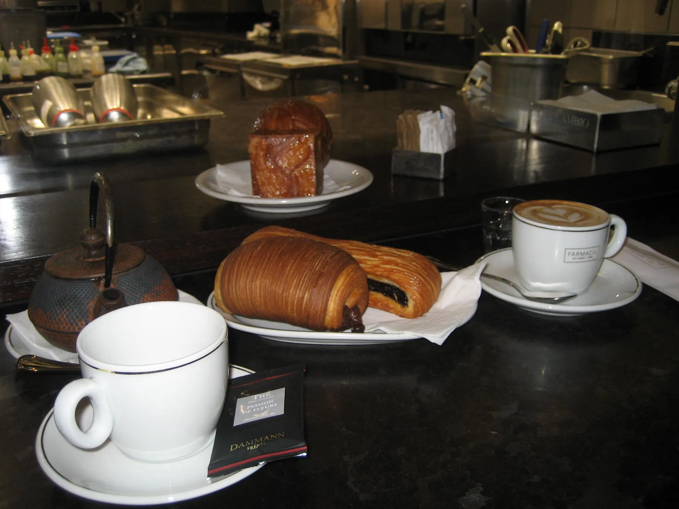 Bancone scuro della Farmacia del Cambio a Torino con un cappuccino, una tazza da tè e piatti con un pain au chocolat tagliato a metà e una brioche quadrata caramellata.