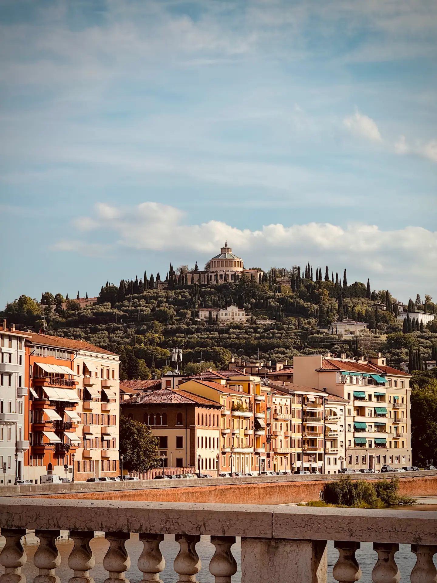 Vista dal Ponte della Vittoria a Verona che mostra una fila di palazzi colorati lungo il fiume Adige e, sullo sfondo, la collina di Castel San Pietro sormontata dal Santuario di Nostra Signora di Lourdes tra i cipressi.